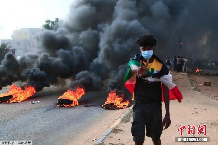 当地时间10月25日，苏丹局势发生突变，过渡政府总理被“绑架”，政府部长被军方抓捕，国家进入紧急状态，大量民众走上街头对军方行动进行抗议，已有7人在抗议活动中死亡，140人受伤。图为一名苏丹男子披着国旗在行走在燃烧的轮胎旁。